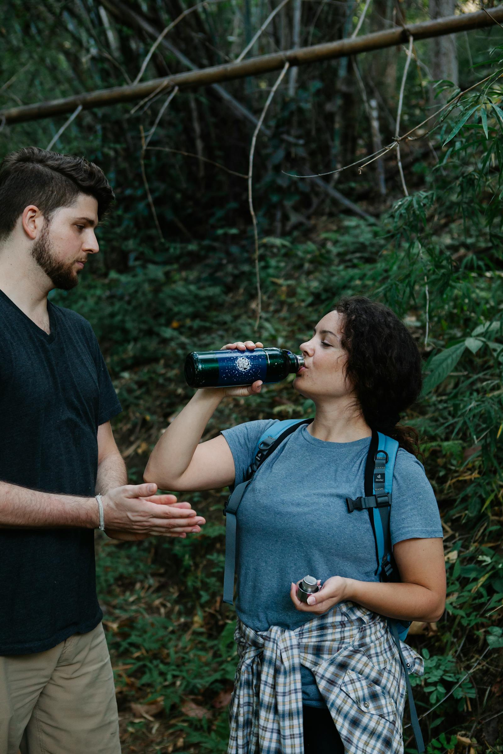 Two hikers in activewear standing in a green outdoor setting while drinking water from a bottle