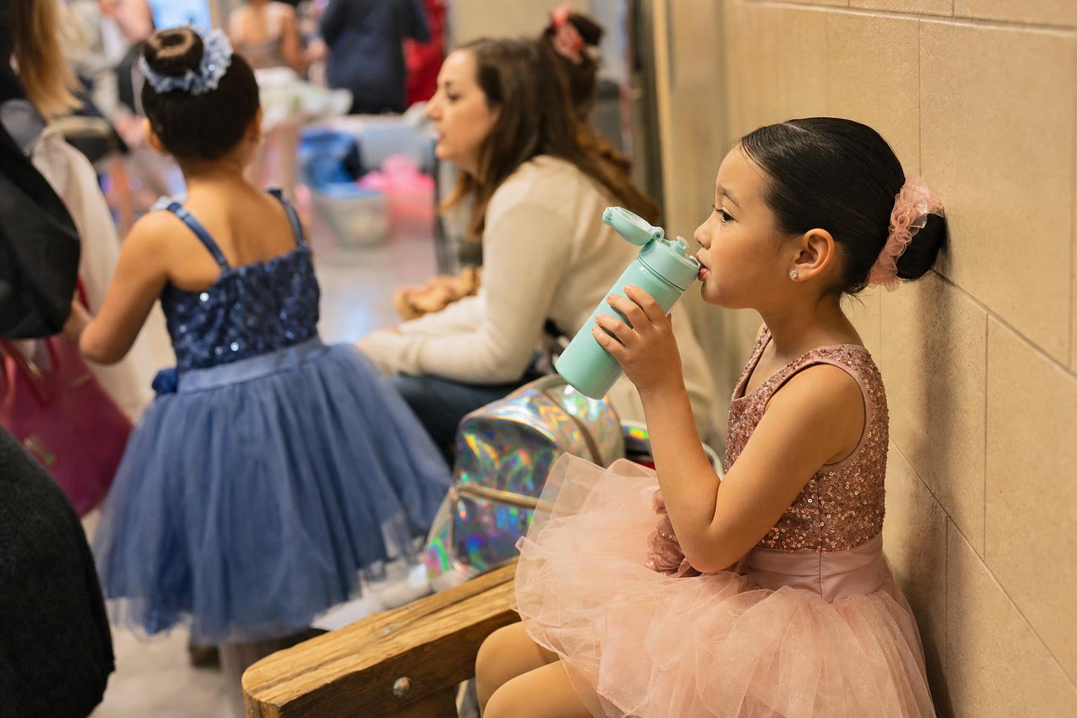 Backstage recital waiting area with children in costumes and a reusable water bottle on a bench