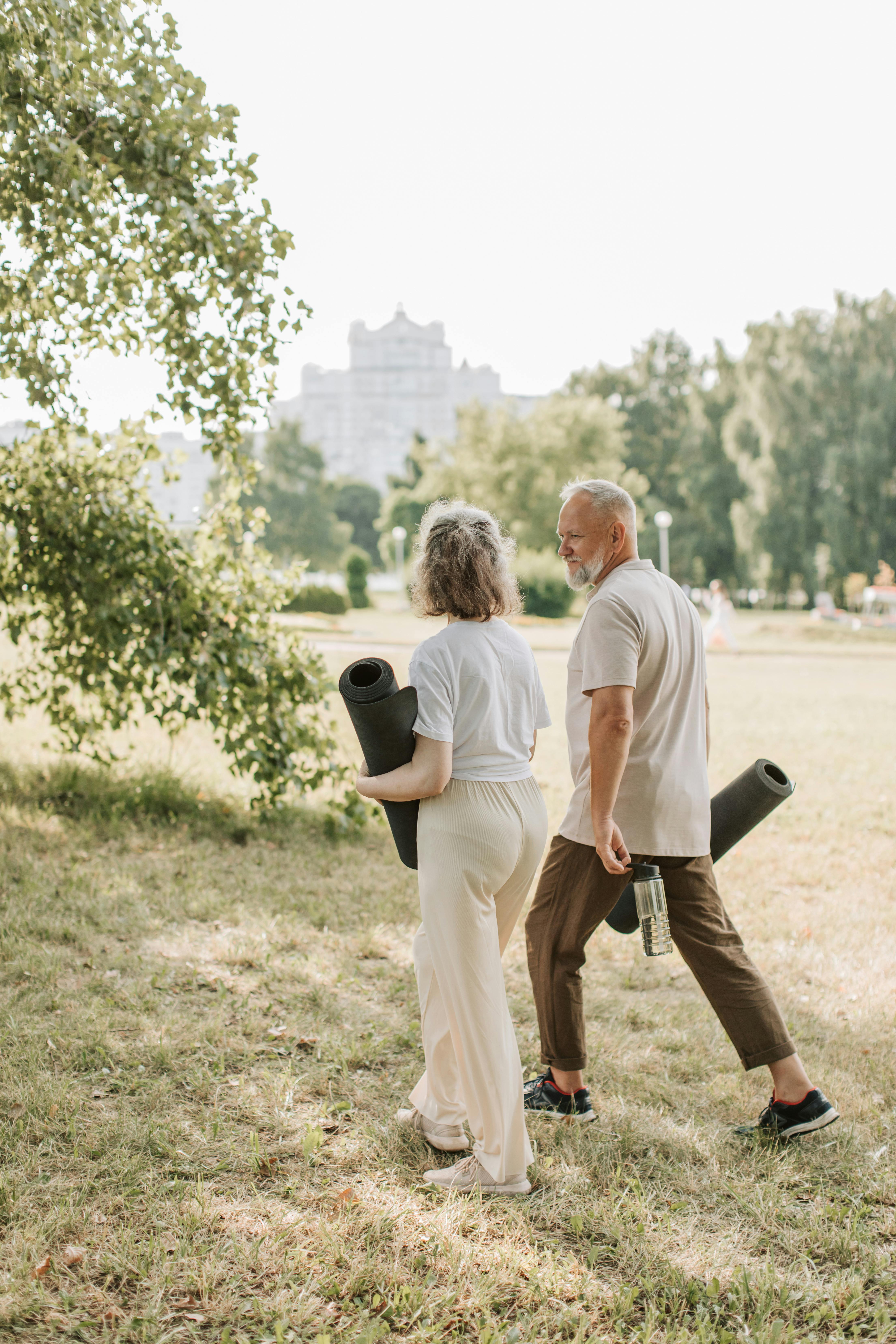 Two adults walking through a sunny park carrying yoga mats and a water bottle before an outdoor class