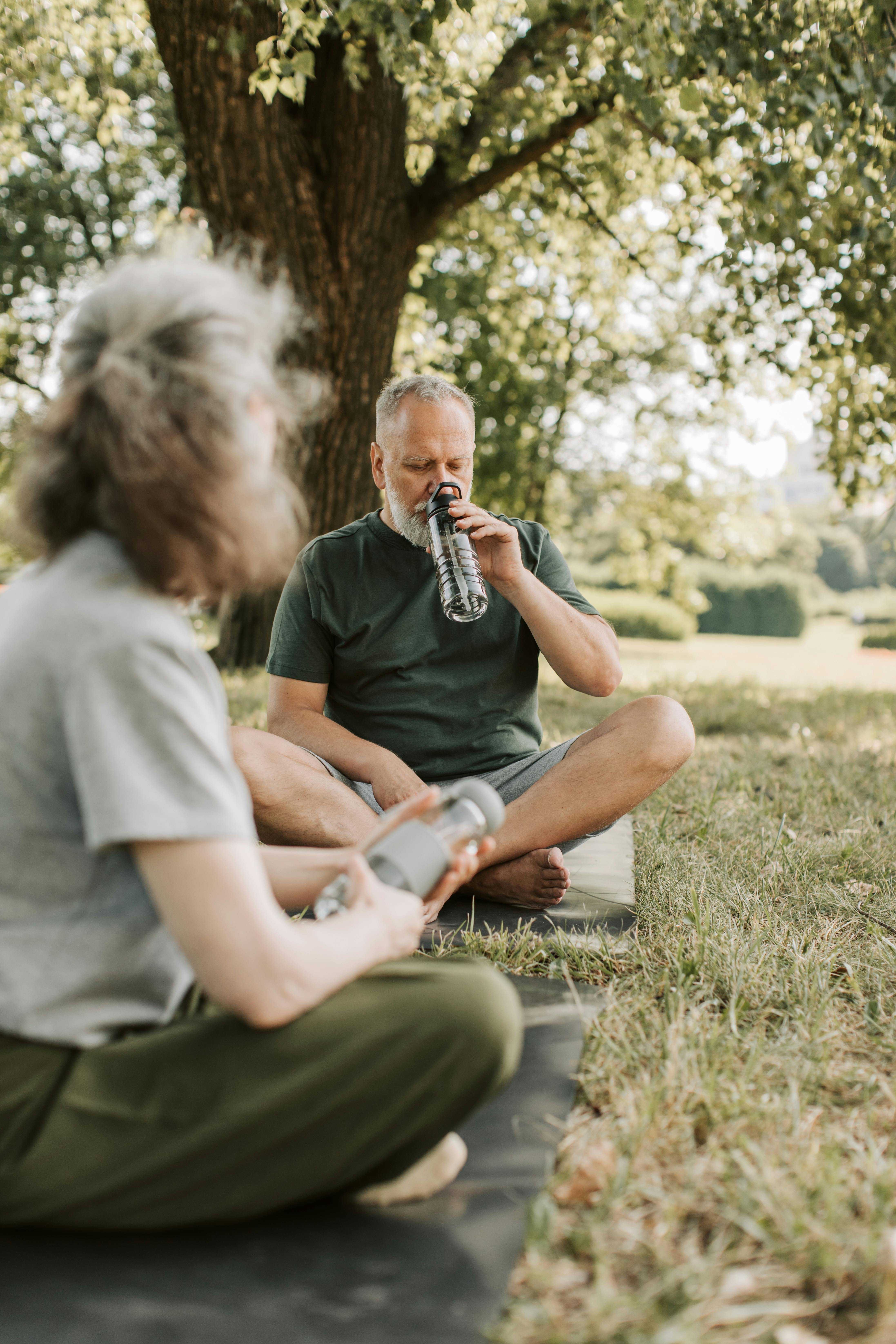 Two adults sitting on yoga mats outdoors while one drinks water after a park workout