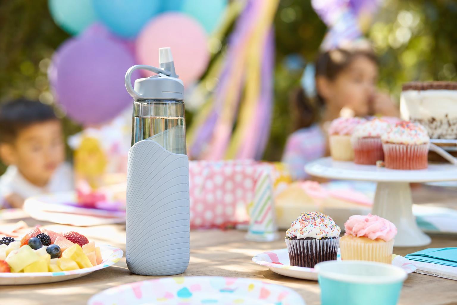 Reusable water bottle on a birthday party table with cupcakes, fruit, and balloons in soft focus behind it